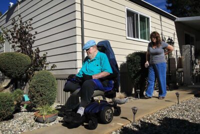 Man in mobile scooter in front yard of home, followed by woman.
