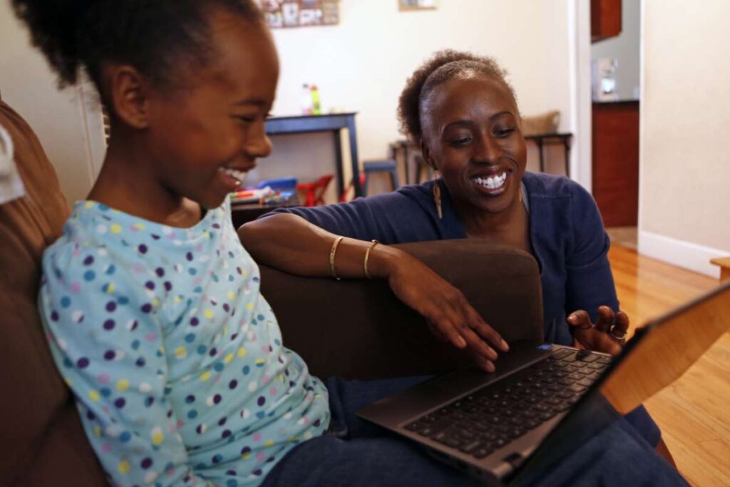 Mother and daughter laugh together while using laptop on couch.