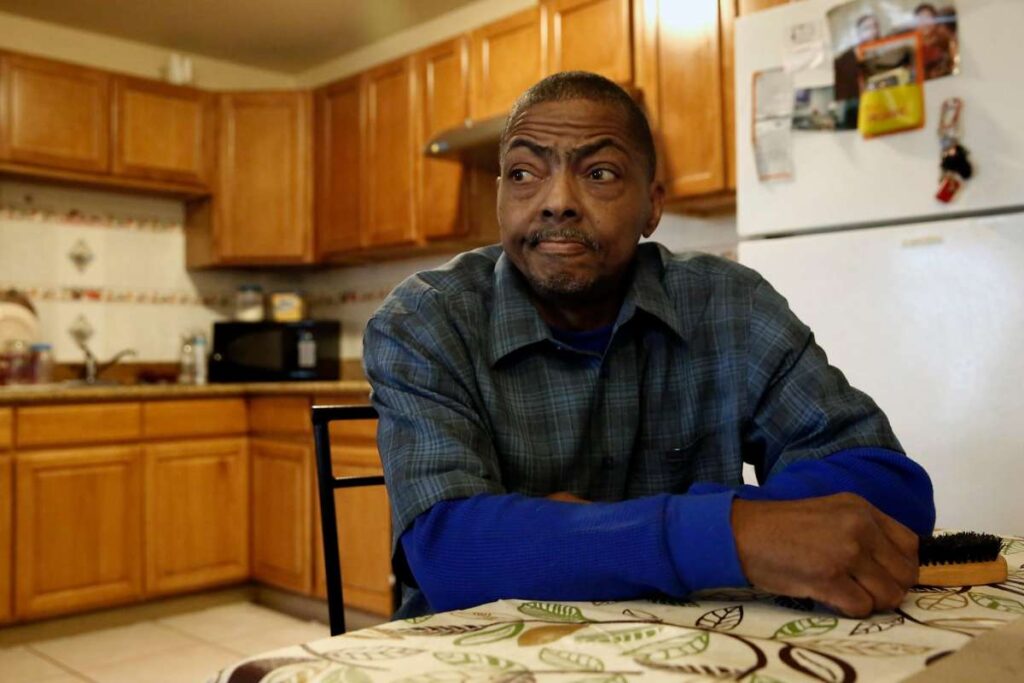 Man sitting in a kitchen with folded arms, appearing contemplative.