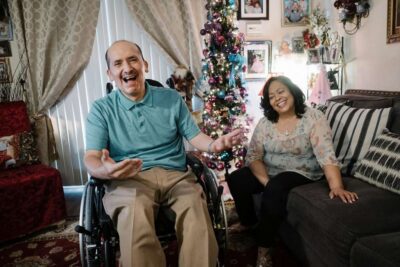 Older couple seated in front of a decorated Christmas tree, smiling warmly, with the man in a wheelchair.