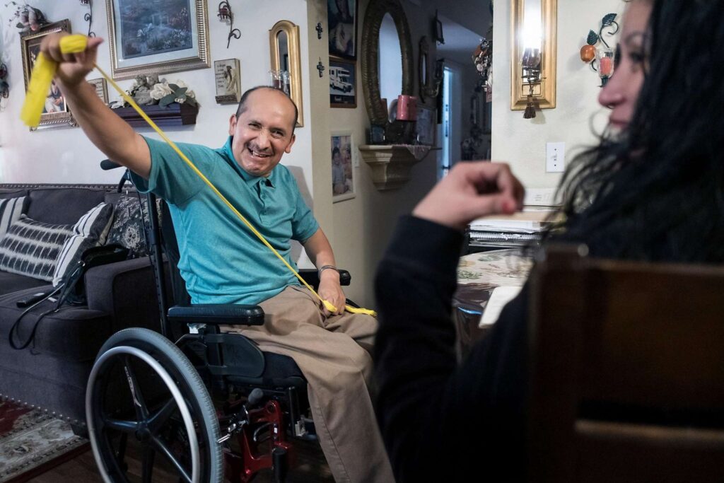 Elderly man in a wheelchair smiling and speaking with a woman in a decorated living room.
