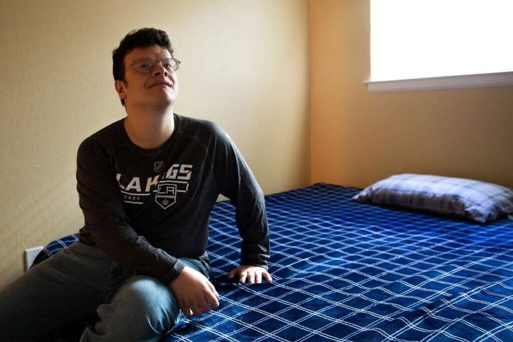 Young man sits on a bed in a room with blue bedding, gazing upward.
