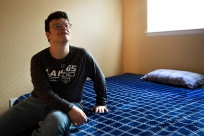 Young man sits on a bed in a room with blue bedding, gazing upward.