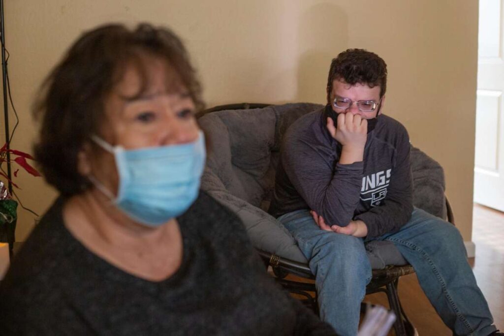 Older woman in a mask sits beside a young man on a couch.