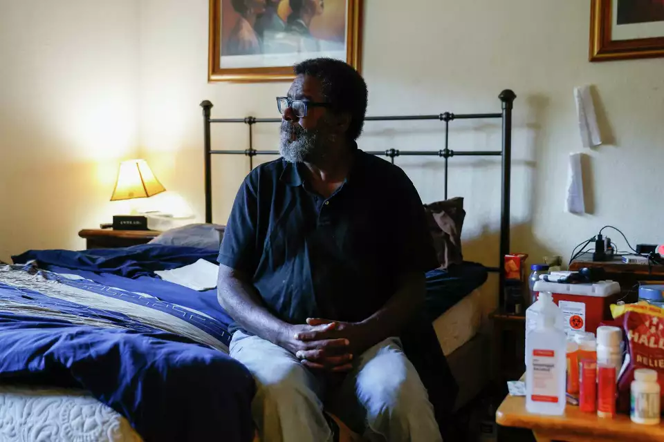 A man sits on the edge of a bed in a dim room—suggesting solitude and contemplation.