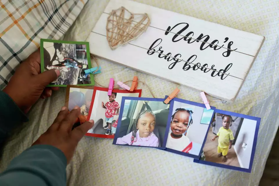 Hand placing photos on a wall display labeled 'Nana’s brag board.'