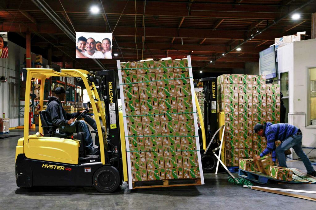 A forklift carries stacks of boxed produce in a food bank warehouse—illustrating community aid.