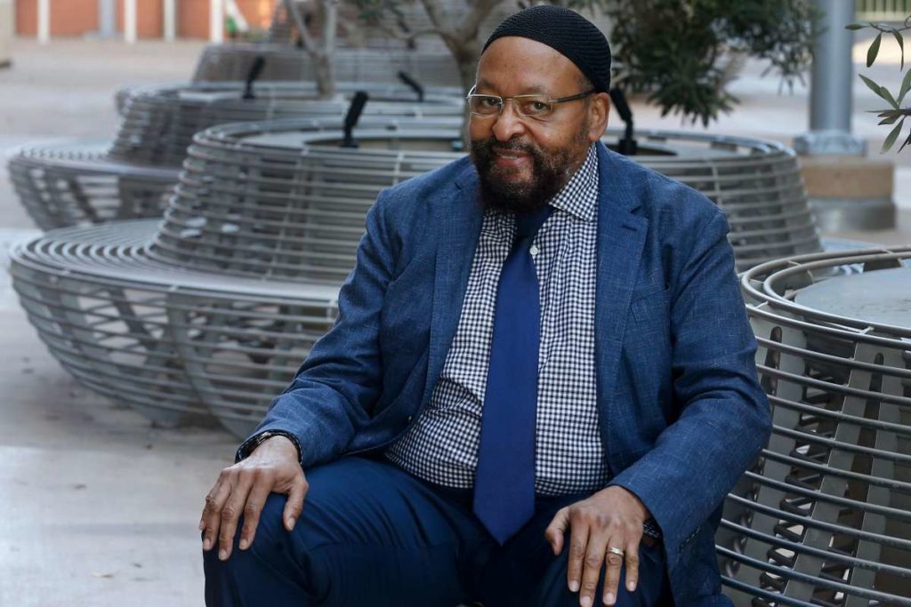 Man in a suit seated outside, smiling.