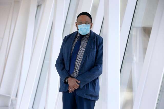 Man wearing a mask stands in a modern office hallway with hands folded.
