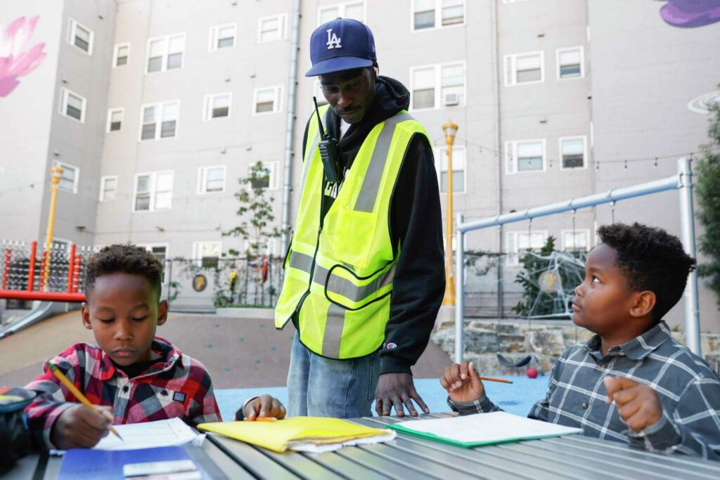 A person in a reflective vest helps children with homework at park—capturing organization and outreach.
