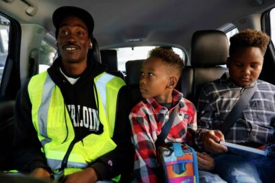 A man wearing safety vest smiles inside a car with two children—showing mentorship and shared purpose.