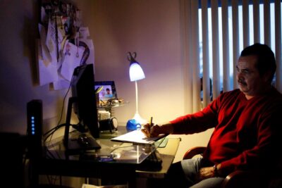 Man sits at computer desk writing in notepad.