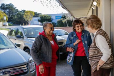 Three women smiling and talking beside a parked car.