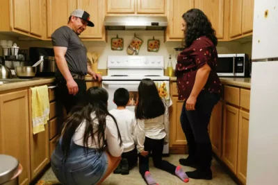 A father and mother prepares food in a kitchen while two children observe—representing learning and nourishment at home.