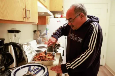 A man in a hoodie prepares food in a modest kitchen, stirring a pot while surrounded by dishes—capturing a moment of care and hospitality.