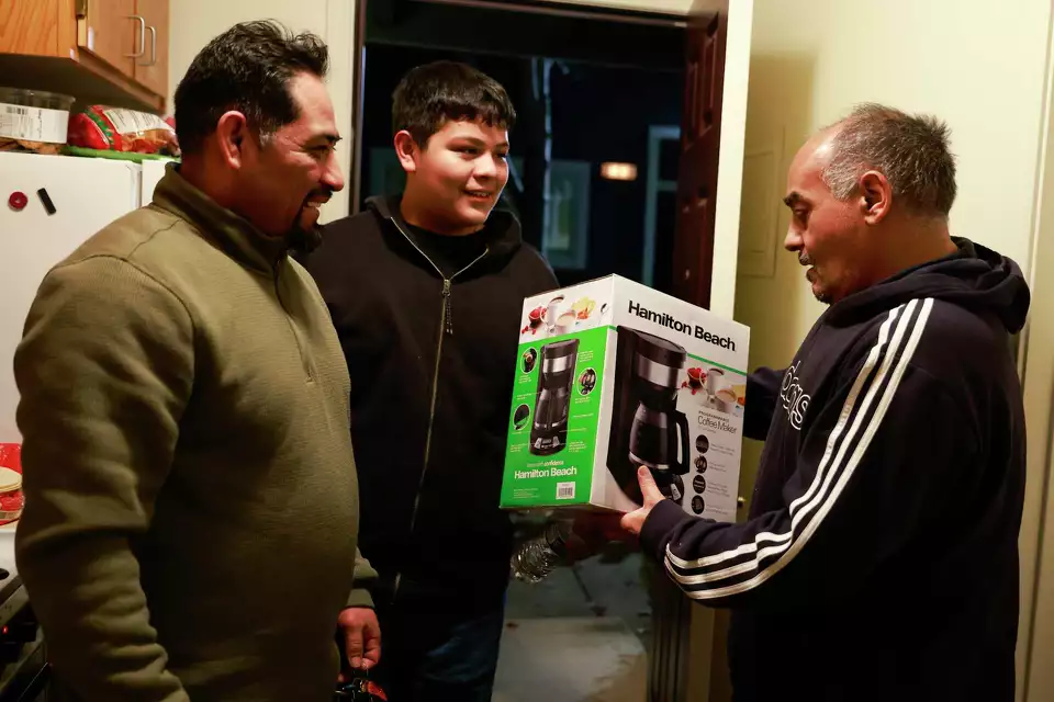 Two men stand in a doorway receiving a boxed household item from another man—showing a gesture of support and giving.