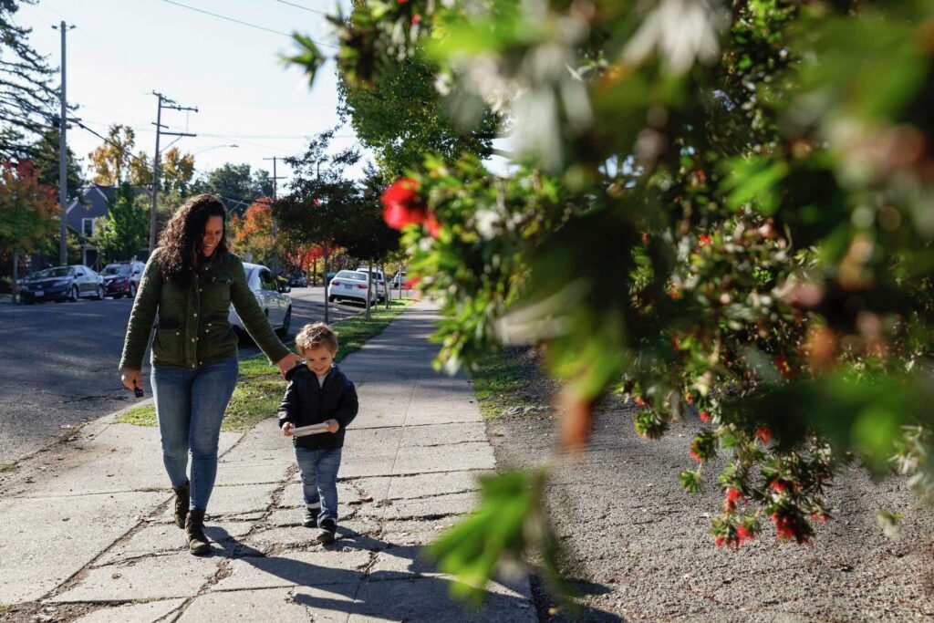 A mother and child walk along a sidewalk near a large tree—highlighting growth and family.
