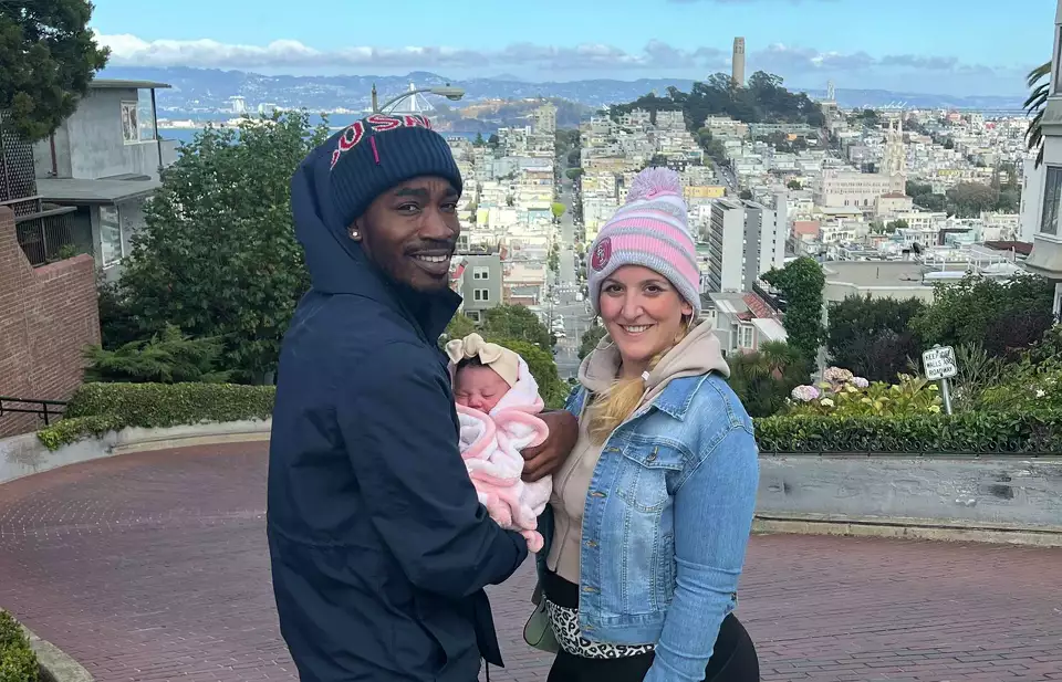 A couple poses with their baby on a scenic hilltop, smiling against a city skyline—illustrating hope and new beginnings.