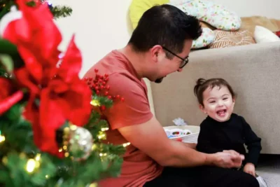 A father and child laugh together near a Christmas tree—sharing joyful family moments.