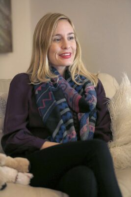 Woman in a scarf sitting on a couch with hands in her lap, looking content.