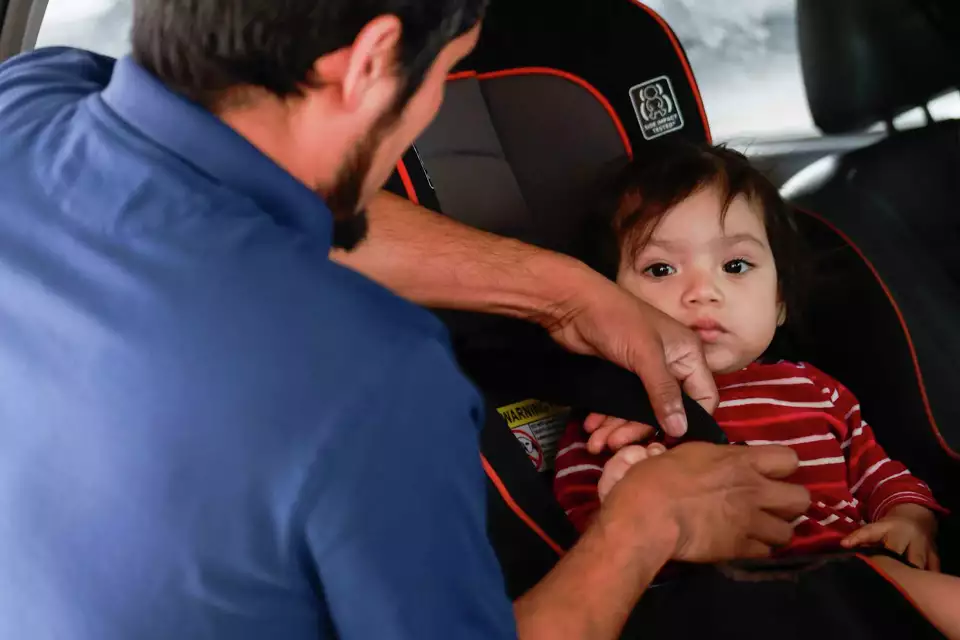 A man carefully secures a baby into a car seat while the child looks on—emphasizing safety, care, and family.