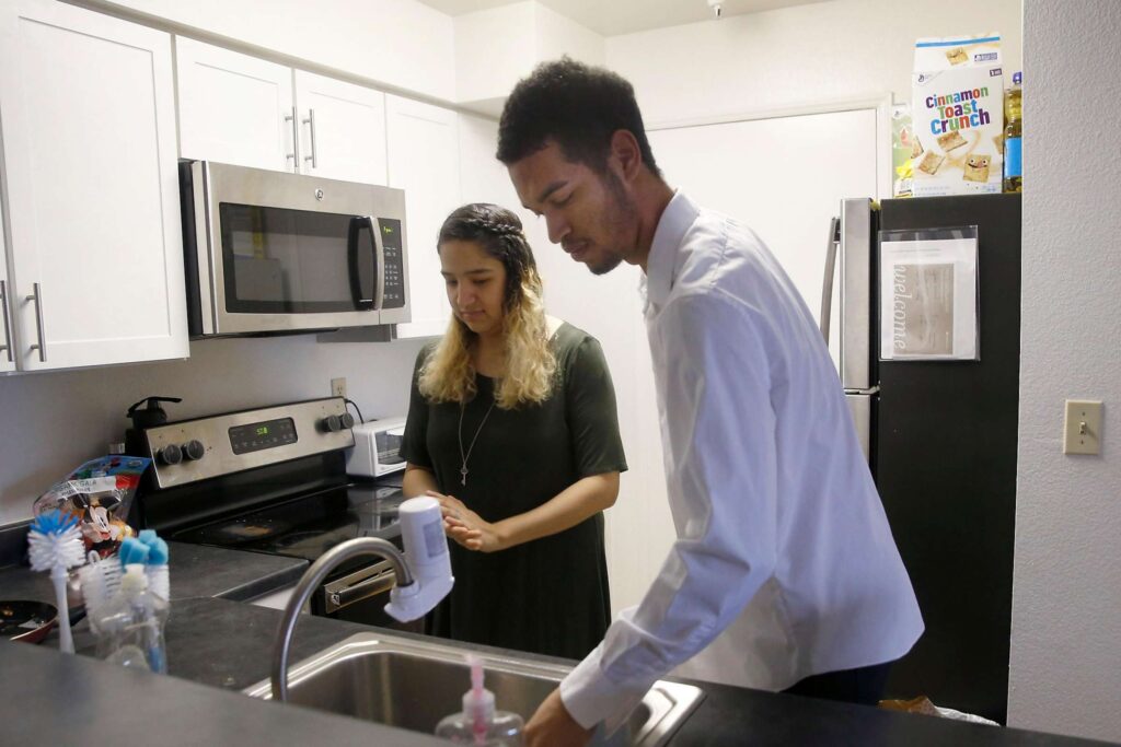 Man washing dishes at the kitchen sink while a woman stands beside him smiling, suggesting shared household responsibilities.
