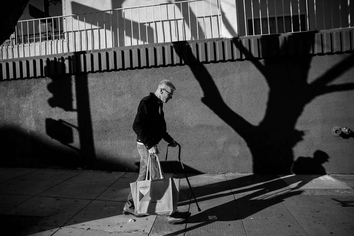 Black and white photo of a man with a cane walking on a sidewalk.