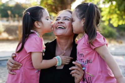 A woman is joyfully hugged by two young girls outdoors—showcasing intergenerational love.