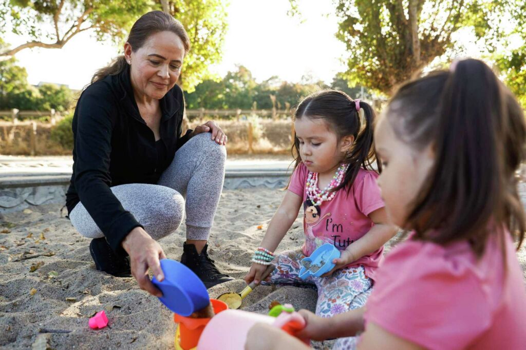 A woman kneels with two girls as they play with sand toys—representing learning and care.