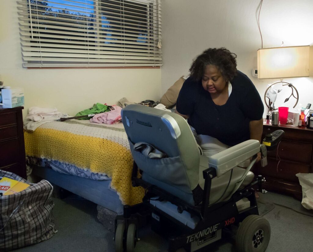 Woman sitting on bed next to mobility scooter in a warmly lit room.