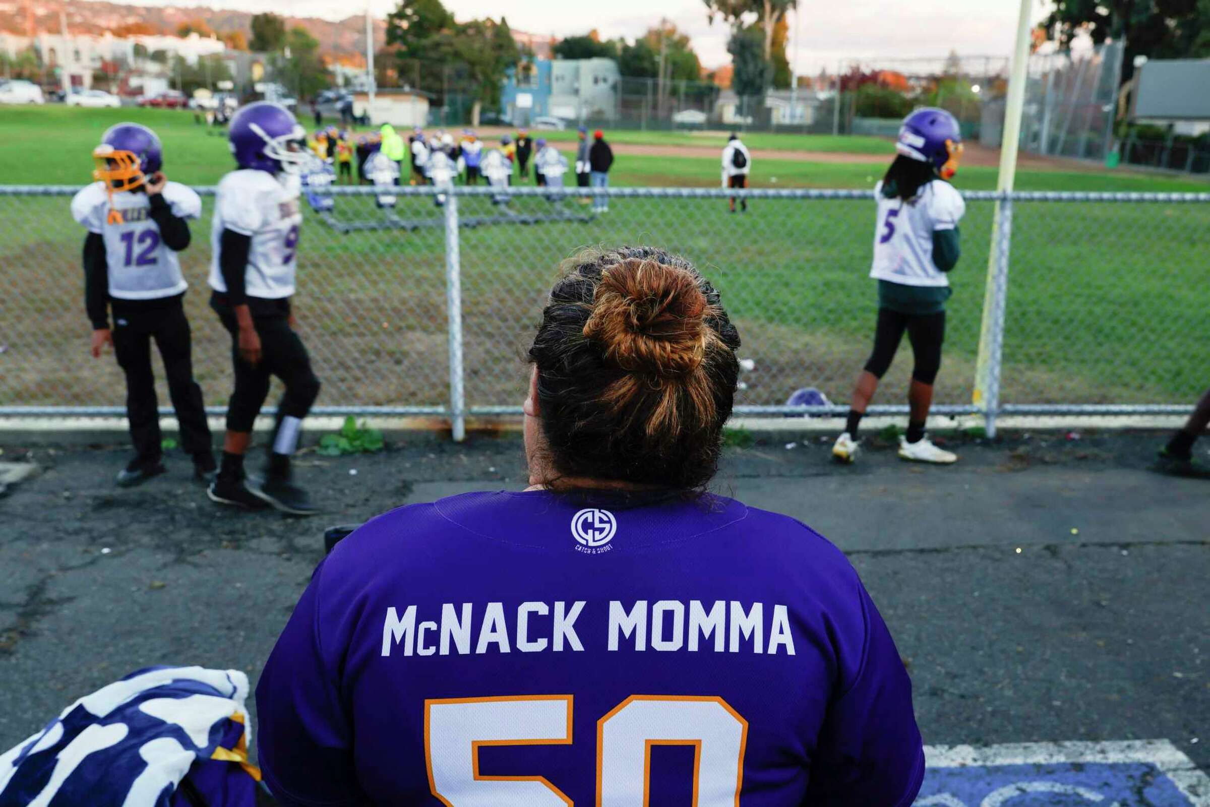 A woman with a jersey labeled “McNack Momma” watches her child's football game—signifying support and pride.