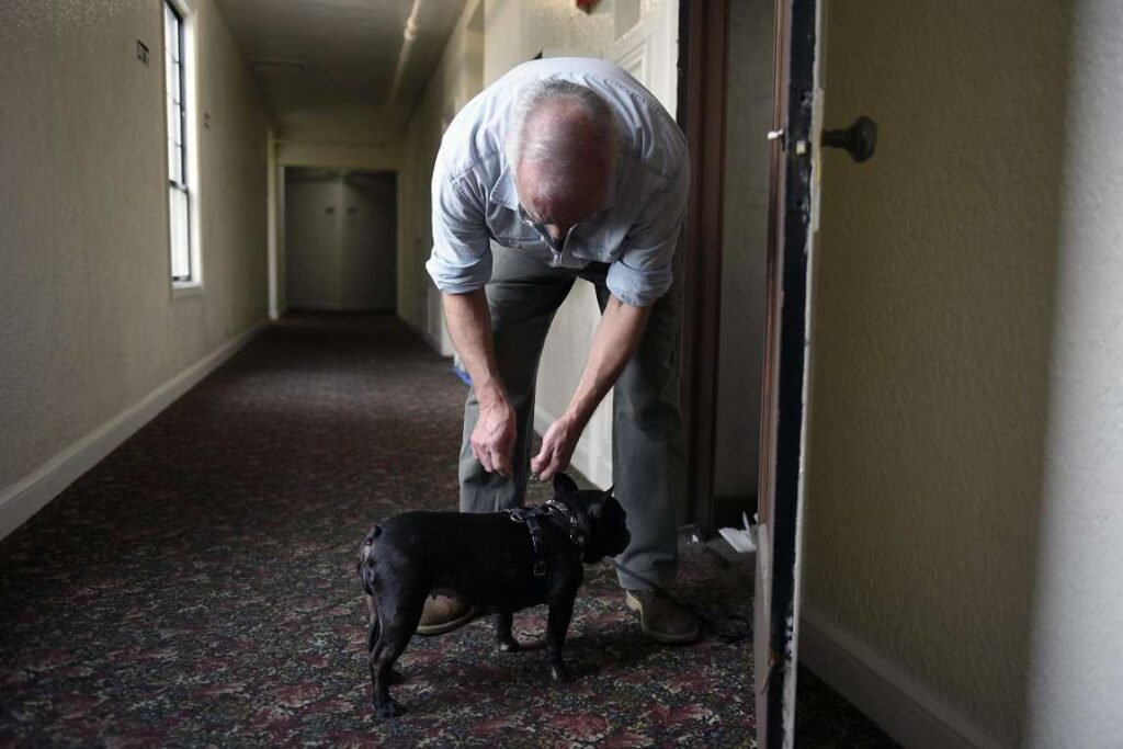 Elderly man bending down to pet a dog in a hallway.