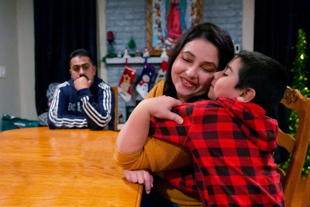 A mother hugs her child at a kitchen table, both smiling brightly—illustrating love and warmth.