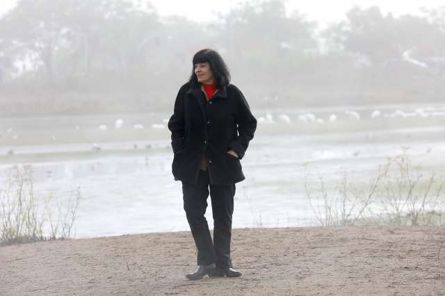 Woman stands alone in front of lake in a winter coat.