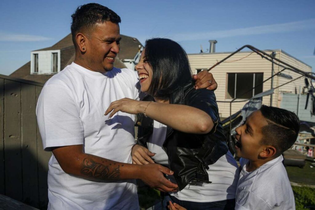 A woman and man embrace joyfully outside their home while a child looks on—symbolizing reunion, love, and family strength.