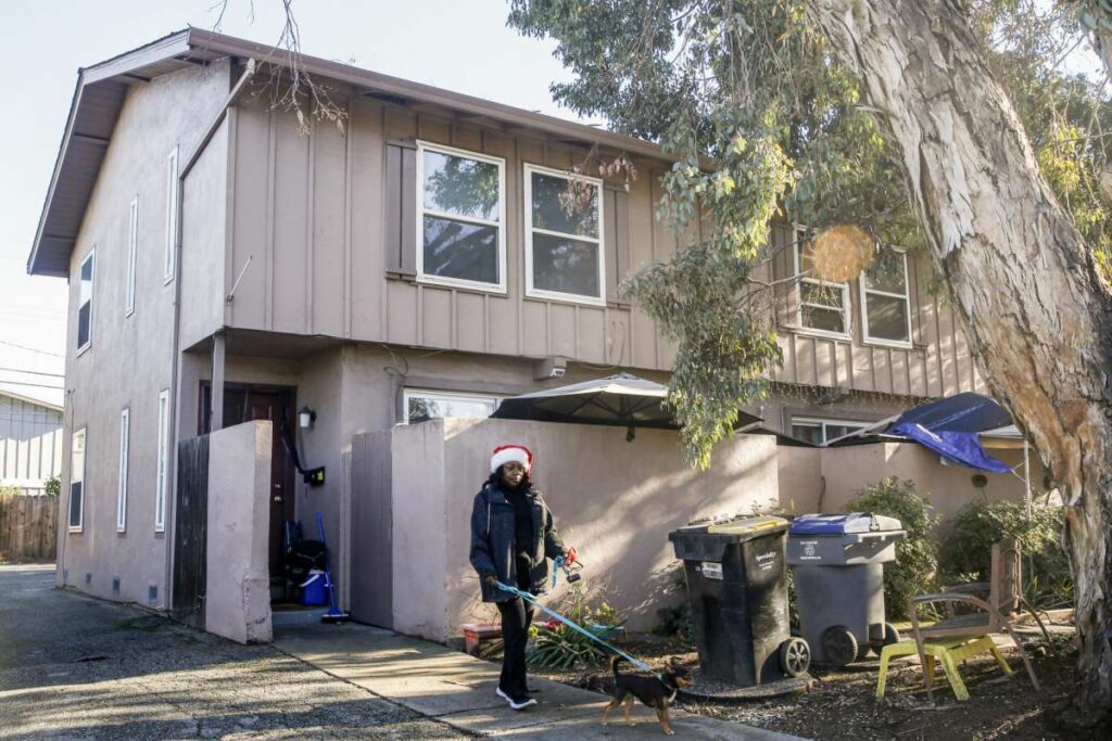 A woman walks outside with small dog beside a home—representing perseverance and daily routine.