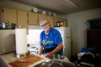 Older man preparing food in a kitchen, smiling at the camera.