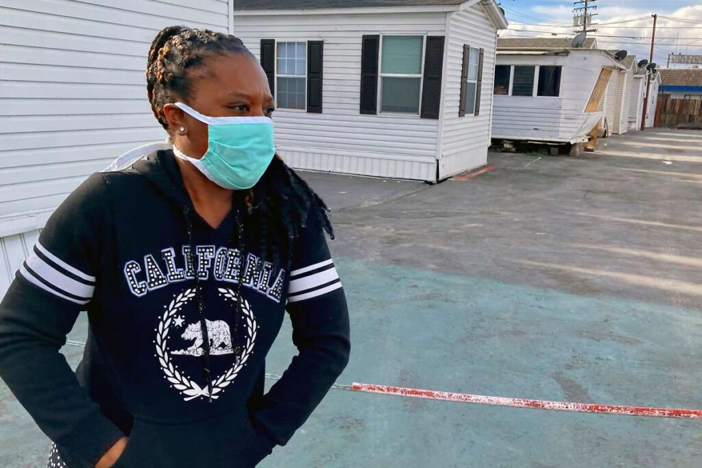 Young woman in a mask stands in a driveway with mobile homes behind her.