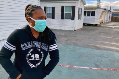 Young woman in a mask stands in a driveway with mobile homes behind her.