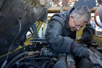 Elderly man in leather jacket repairs car engine.