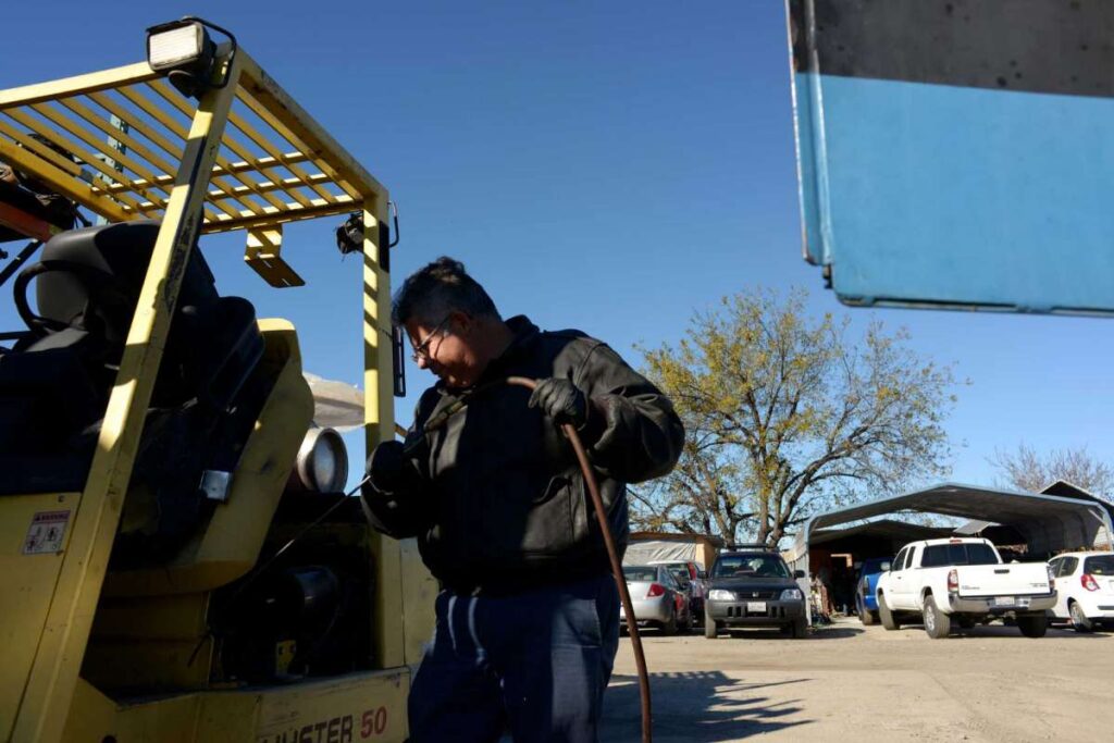 Man in leather jacket repairs a forklift outdoors.