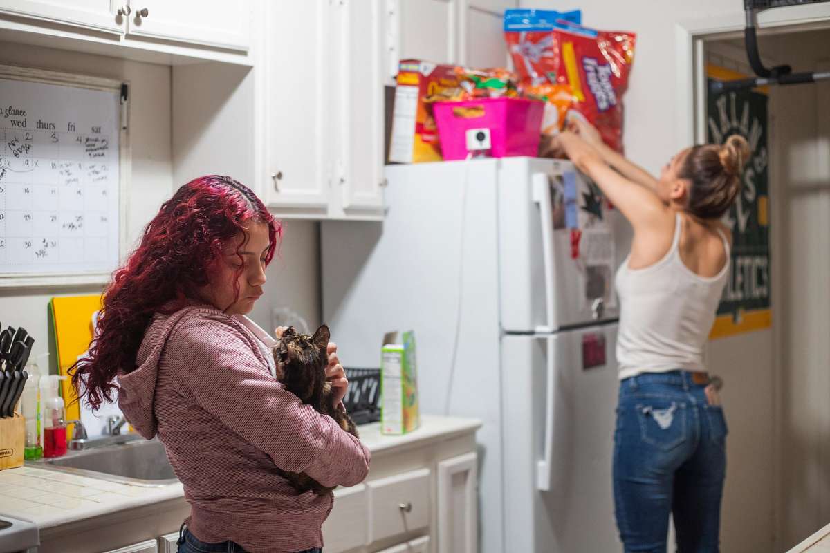 Two women interact in a kitchen as one places food on the counter and other holds her cat.