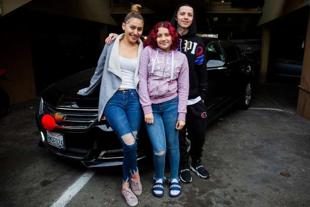 Young family of three stand smiling together on a driveway.
