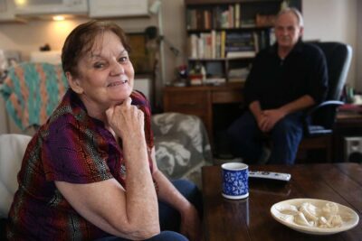 Woman smiles and poses for photo on couch with husband in background.