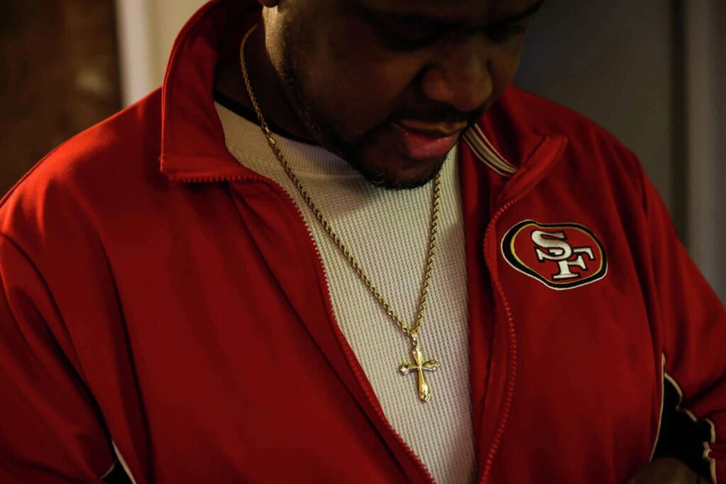 A man in a red sports jacket and cross necklace looks down with emotion—evoking strength and vulnerability.