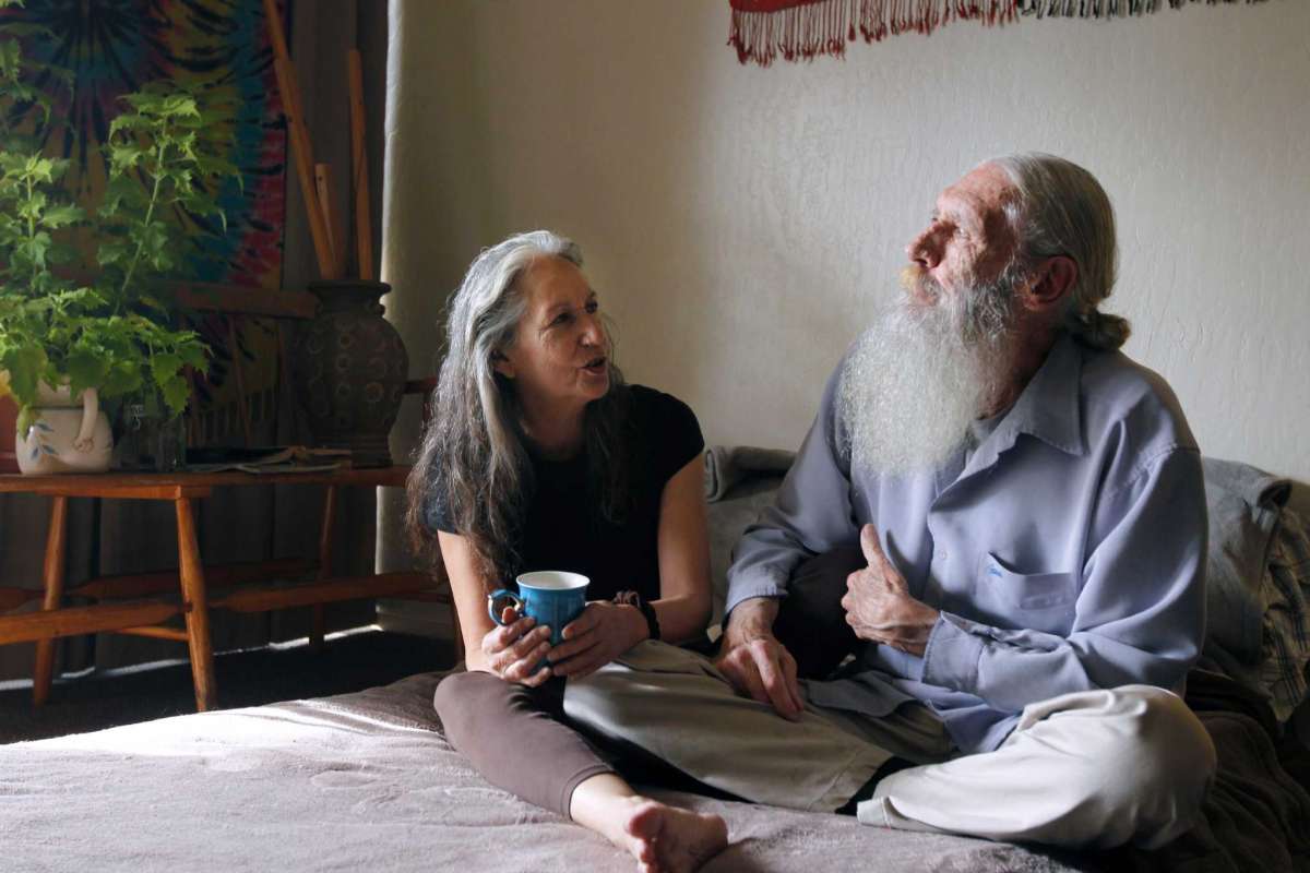 Elderly couple sit on bed together with cup of coffee.