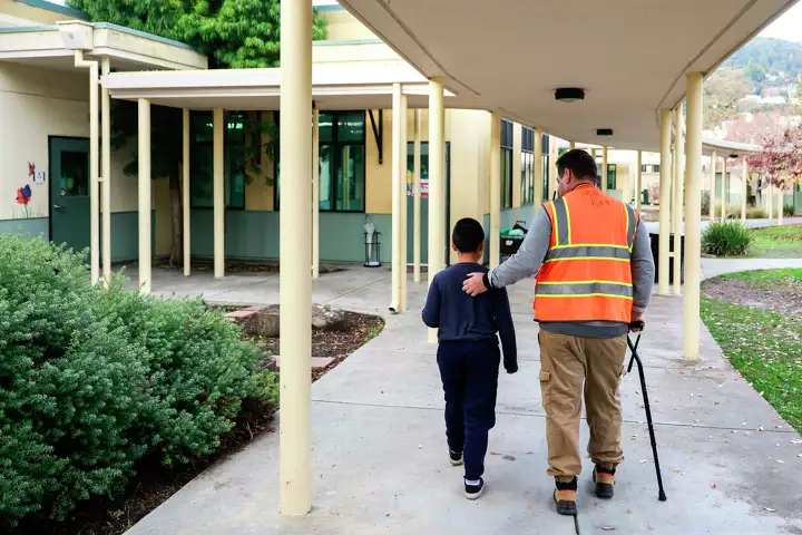 A staff member with walking cane supportingly guides a student down a school hallway—portraying guidance and support.