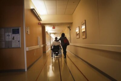 Man in wheelchair and woman making way down hallway of hospital.