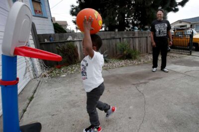 Child plays with basketball on driveway while father watches.