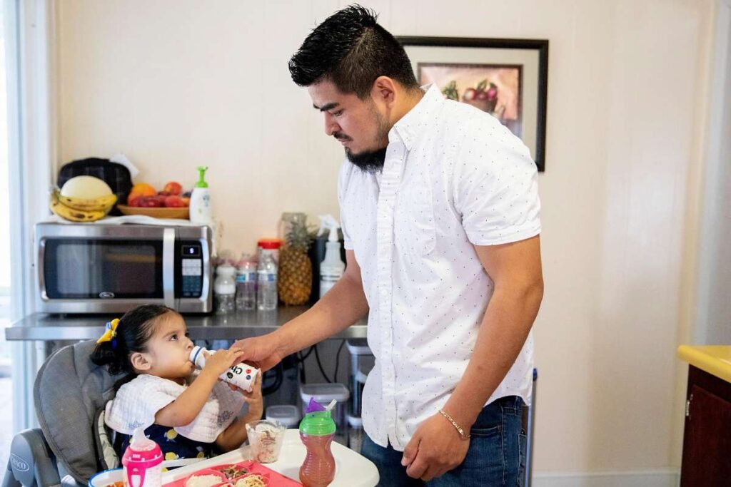Father serves young daughter food in high chair.